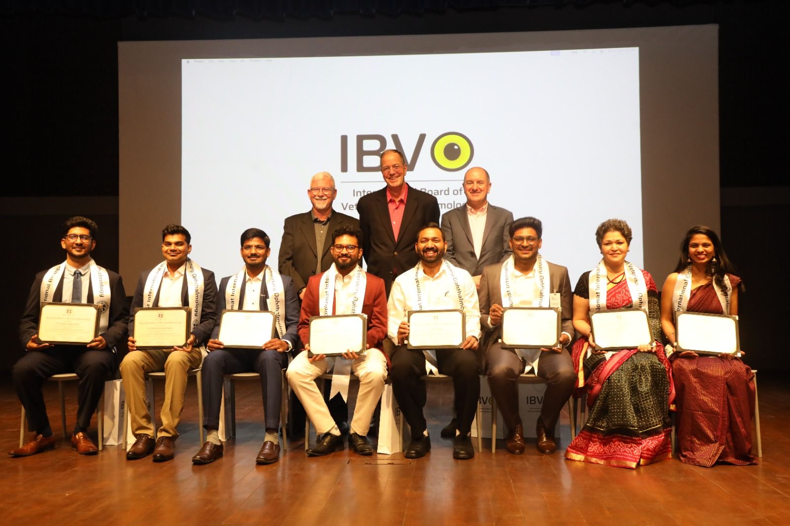 The eight new IBVO Diplomates seated on stage holding their certificates, the three founders standing behind them and the IBVO logo on the screen